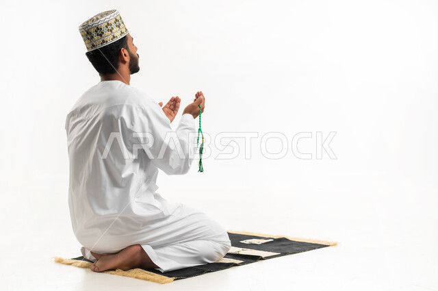 الإكثار من ذكر الله تعالى من خلال الإستغفار والدعاء, الإجتهاد في العباده والتقرب الى الله في شهر رمضان المبارك, مفهوم التسبيح والتهليل، بورتريه لشاب عربي خليجي عماني يرتدي الدشداشة والكمه يجلس على سجادة الصلاة يمسك السبحه ويرفع يديه تضرع بالدعاء, خلفيه بيضاء