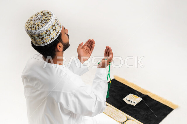 الإجتهاد في العباده والتقرب الى الله في شهر رمضان المبارك, مفهوم التسبيح والتهليل، بورتريه مقرب لشاب عربي خليجي عماني يرتدي الدشداشة والكمه يجلس على سجادة الصلاة يمسك السبحه ويرفع يديه تضرع بالدعاء, الإكثار من ذكر الله تعالى من خلال الإستغفار والدعاء, خلفيه بيضاء