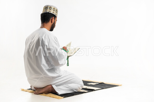 الإكثار من ذكر الله تعالى، تلاوة وقراءة القرآن الكريم، بورتريه من الخلف لشاب عربي خليجي عماني يرتدي الدشداشة والكمه يجلس على سجادة الصلاة ويحمل بيده المصحف الشريف، الاجتهاد في العبادة، التقرب الى الله في شهر رمضان، الشعور بالخشوع والطمأنينة، خلفية بيضاء