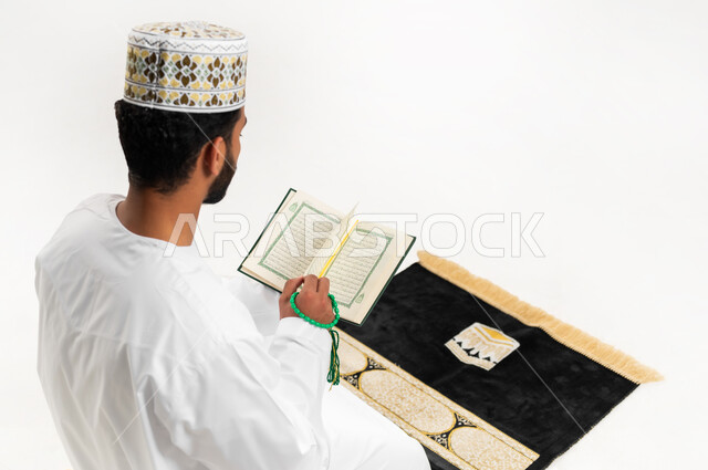 تلاوة وقراءة القرآن الكريم، بورتريه مقرب من الخلف لشاب عربي خليجي عماني يرتدي الدشداشة والكمه يجلس على سجادة الصلاة ويحمل بيده المصحف الشريف، الإكثار من ذكر الله تعالى، الاجتهاد في العبادة، التقرب الى الله في شهر رمضان، الشعور بالخشوع والطمأنينة، خلفية بيضاء