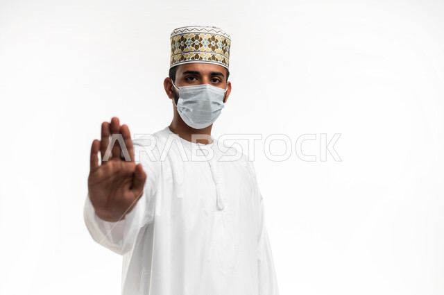 Wearing a mask to prevent the Corona virus Covid-19, maintaining distances and avoiding superficial contact, close-up portrait of a young Arab Gulf Omani man wearing a dishdasha and a sleeve looking at the camera and raising his hand to avoid shaking hands and not letting others approach him, adhering to preventive measures, white background