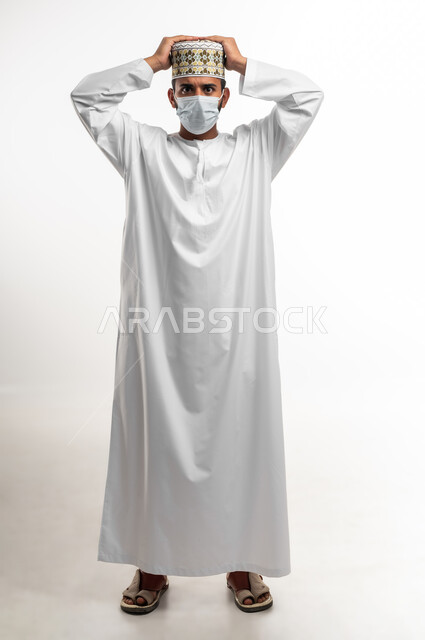 المعاناة من الصداع وألم الرأس ، تعابير الاستياء والضجر من المرض، اتباع الاجراءات الاحترازية للوقاية من فايروس كورونا، بورتريه لشاب عربي خليجي عماني يرتدي الدشداشة والكمه يضع الكمامة يرفع يديه على رأسه بايماءات الارهاق، الطول الكامل للجسم، خلفية بيضاء