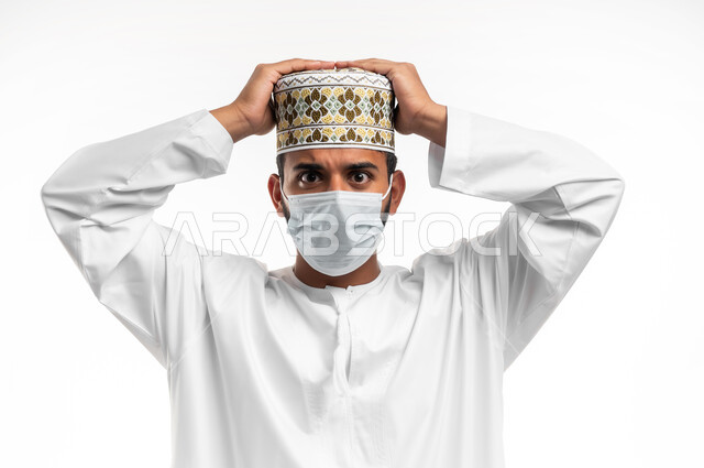 تعابير الاستياء والضجر من المرض، اتباع الاجراءات الاحترازية للوقاية من فايروس كورونا، الصداع وألم الرأس والمعاناة من آلام الشقيقة، بورتريه مقرب لشاب عربي خليجي عماني يرتدي الدشداشة والكمه يضع الكمامة يرفع يديه على رأسه بايماءات الارهاق، خلفية بيضاء
