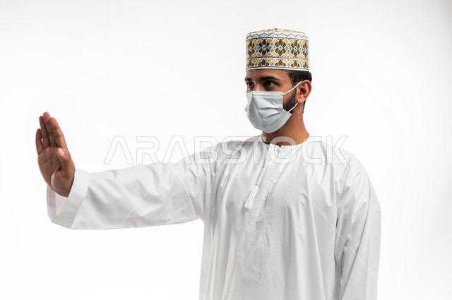 الحفاظ على المسافات وعدم الملامسة السطحية، بورتريه مقرب لشاب عربي خليجي عماني يرتدي الدشداشة والكمه يرفع يده لتجنب المصافحة وعدم اقتراب الآخرين منه، وضع القناع الطبي للوقاية من فايروس كورونا كوفيد-19، الالتزام بالإجراءات والقواعد الوقائية، خلفية بيضاء
