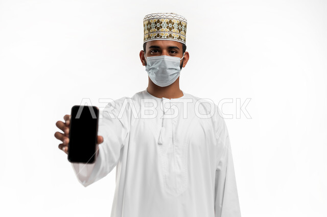 ارتداء الكمامة لإجراءات الوقاية من فايروس كورونا, التوعيه من مخاطر كوفيد 19، بورتريه مقرب لشاب عربي خليجي عماني يرتدي الدشداشة والكمه يعرض شاشة سوداء  فارغه على الهاتف المحمول، احترام قوانين وقواعد وزارة الصحة، خلفيه بيضاء