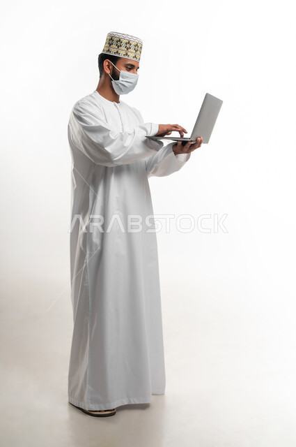 Using a laptop to complete tasks and projects remotely, a portrait of a young Arab Gulf Omani man wearing a dishdasha and a sleeve, wearing a medical mask to protect against the Corona virus Covid-19, respecting the laws and rules of the Ministry of Health, full length of the body, white cell