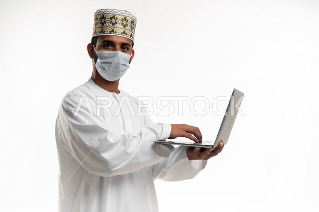 احترام قوانين وقواعد وزارة الصحة، استخدام جهاز الحاسوب المحمول لإنجاز المهام والمشاريع عن بعد، النظر الى الكاميرا بتعابير الرضا, بورتريه مقرب لشاب عربي خليجي عماني يرتدي الدشداشة والكمه يضع القناع الطبي للوقاية من فايروس كورونا كوفيد-19، خلفية بيضاء