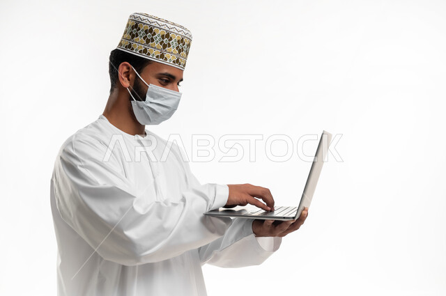 استخدام جهاز الحاسوب المحمول لإنجاز المهام والمشاريع عن بعد، احترام قوانين وقواعد وزارة الصحة، بورتريه مقرب لشاب عربي خليجي عماني يرتدي الدشداشة والكمه يضع القناع الطبي للوقاية من فايروس كورونا كوفيد-19، خلفية بيضاء