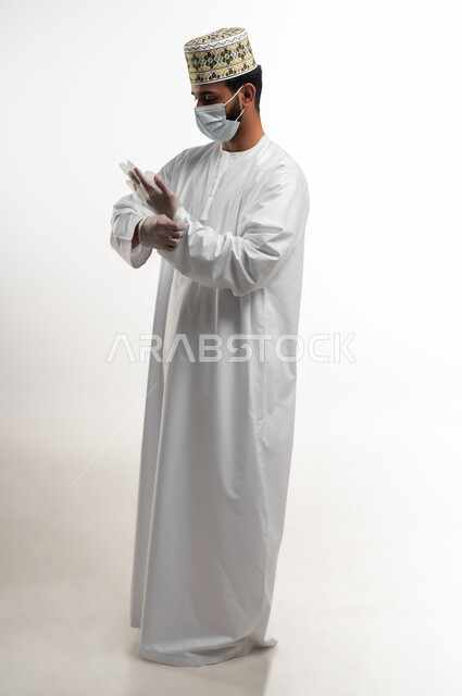احترام قوانين وقواعد وزارة الصحة لتجنب الاصابة من كوفيد 19، اتباع الاحترازات الوقائية من فايروس كورونا، بورتريه لشاب عربي خليجي عماني يرتدي الدشداشة والكمه يضع الكمامة والقفازات للحفاظ على السلامة, صورة الجسم بالكامل، خلفية بيضاء