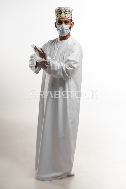اتباع الاحترازات الوقائية من فايروس كورونا، احترام قوانين وقواعد وزارة الصحة لتجنب الاصابة من كوفيد 19، بورتريه لشاب عربي خليجي عماني يرتدي الدشداشة والكمه يضع الكمامة والقفازات للحفاظ على السلامة, صورة الجسم بالكامل، خلفية بيضاء