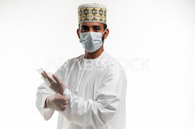 النظر الى الكاميرا بايماءات الرضا, احترام قوانين وقواعد وزارة الصحة لتجنب الاصابة من كوفيد 19، اتباع الاحترازات الوقائية من فايروس كورونا، بورتريه مقرب لشاب عربي خليجي عماني يرتدي الدشداشة والكمه يضع الكمامة والقفازات للحفاظ على السلامة, خلفية بيضاء