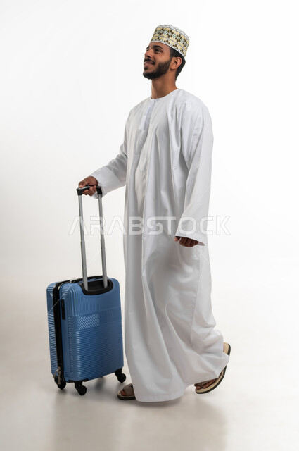 The concept of tourism and travel, spending enjoyable times on vacation, a portrait of a young Arab Gulf Omani man wearing a dishdasha and a cuff and wearing a headphone, holding a suitcase in his hand, looking at something with expressions of enthusiasm and pleasure, preparing for the trip, body full length, white background.