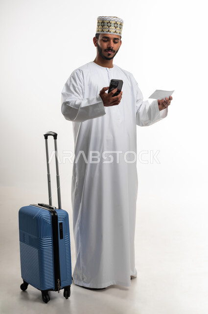 إرسال رسالة نصية عبر الجوال للتأكد من موعد الحجز, قضاء أوقات ممتعه في السياحة وقت العطلة الصيفية, بورتريه  لشاب عربي خليجي عماني يرتدي الدشداشه والكمه يقوم باستخدام الهاتف المحمول وبجانبه حقيبة السفر, الاستعداد لوقت الرحلة، الطول الكامل للجسم, خلفية بيضاء