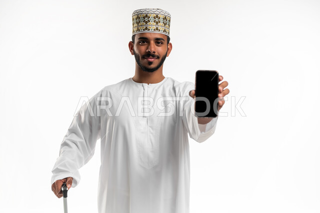 الاستعداد لرحلة ترفيهية ممتعة, عرض شاشة سوداء فارغة على الموبايل, قضاء أوقات العطلة الصيفية في السياحة والسفر, بورتريه  مقرب لشاب عربي خليجي عماني يرتدي الدشداشه والكمه يقوم باستخدام الهاتف المحمول يمسك بيده حقيبة السفر, خلفية بيضاء