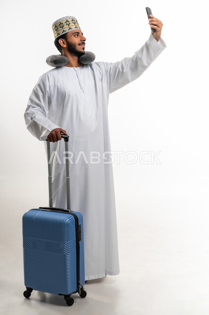 التقاط السيلفي لتوثيق اللحظات التذكارية أثناء الرحلة, مفهوم الاسترخاء والاستراحة, بورتريه  لشاب عربي خليجي عماني يرتدي الدشداشه والكمه يضع وسادة النوم على رقبته يمسك بيده الهاتف المحمول وحقيبة السفر, صورة الجسم بالطول الكامل,  خلفية بيضاء