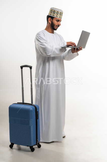 Completing remote work tasks, preparing for an enjoyable travel trip, using advanced tourism devices, technologies and applications, portrait of an Arab Gulf Omani young man wearing a dishdasha and a cuff holding a travel bag and laptop, gestures of integration and focus into the laptop, white background