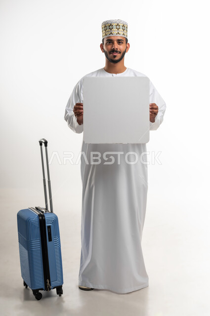 الترويج على لوح إعلاني فارغ، النظر الى الكاميرا بإيماءات السرور، الاستعداد لرحلة ترفيهية ممتعة، بورتريه  لشاب عربي خليجي عماني يرتدي الدشداشه والكمه يحمل ورقة  بيضاء كبيرة وبجانبه حقيبة السفر, مفهوم السياحة والسفر، الطول الكامل للجسم, خلفية بيضاء