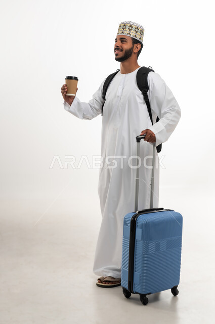 الاستعداد لوقت الرحلة، مفهوم السياحة والسفر، قضاء أوقات ممتعة في الإجازة, بورتريه لشاب عربي خليجي عماني يرتدي الدشداشة و الكمه يمسك كأس القهوة وحقيبة السفر بيده, النظر الى شيء ما بتعابير الحماس والسرور, الجسم بالطول الكامل, خلفية بيضاء