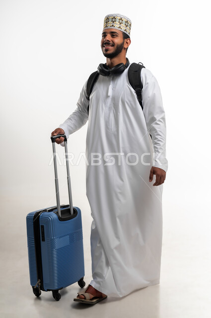 The concept of tourism and travel, spending enjoyable times on vacation, a portrait of a young Arab Gulf Omani man wearing a dishdasha and a cuff and wearing a headphone, holding a suitcase in his hand, looking at something with expressions of enthusiasm and pleasure, preparing for the trip, body full length, white background.