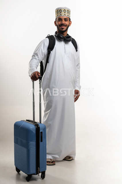 قضاء أوقات ممتعة في الإجازة, بورتريه لشاب عربي خليجي عماني يرتدي الدشداشة و الكمه ويضع سماعة الهيدفون يمسك حقيبة السفر بيده, النظر الى الكاميرا بتعابير الحماس والسرور, مفهوم السياحة والسفر، الاستعداد لوقت الرحلة، الجسم بالطول الكامل, خلفية بيضاء