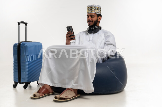 تصفح تطبيقات الهاتف المحمول، التحدث والدردشة مع الاصدقاء عبر الانترنت، استخدام التقنيات الحديثة المتطورة، بورتريه  لشاب عربي خليجي عماني يرتدي الدشداشه والكمه يضع سماعة الهيدفون يجلس على كرسي مريح يقوم بمتابعة اخبار وسائل التواصل وبجانبه حقيبة السفر، خلفية بيضاء