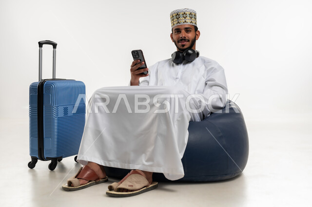 النظر الى الكاميرا بايماءات السرور، تصفح تطبيقات الهاتف المحمول، التحدث والدردشة مع الاصدقاء عبر الانترنت، بورتريه  لشاب عربي خليجي عماني يرتدي الدشداشه والكمه يجلس على كرسي مريح يقوم بمتابعة اخبار وسائل التواصل الاجتماعي وبجانبه حقيبة السفر، خلفية بيضاء