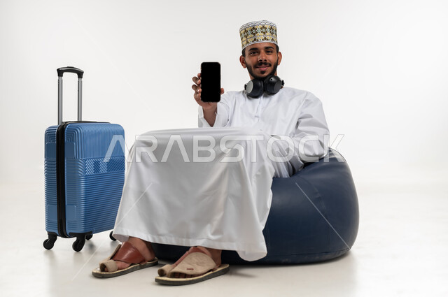 Using technology and advanced modern techniques, looking at the camera with expressions of happiness and pleasure, a portrait of a young Arab Gulf Omani man wearing a dishdasha and a sleeve and wearing a headphone, sitting on a comfortable chair with a suitcase next to him, showing a blank black screen on a mobile phone, white background.