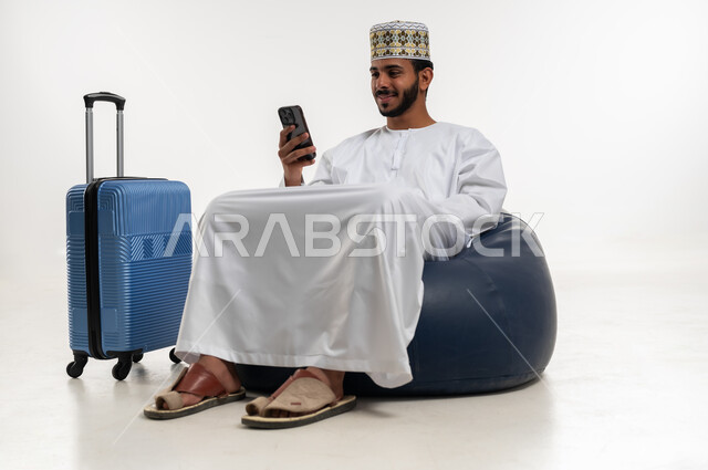 تصفح تطبيقات الهاتف المحمول، التحدث والدردشة مع الاصدقاء عبر الانترنت، استخدام التقنيات الحديثة المتطورة، بورتريه  لشاب عربي خليجي عماني يرتدي الدشداشه والكمه يجلس على كرسي مريح يقوم بمتابعة اخبار وسائل التواصل الاجتماعي وبجانبه حقيبة السفر، خلفية بيضاء