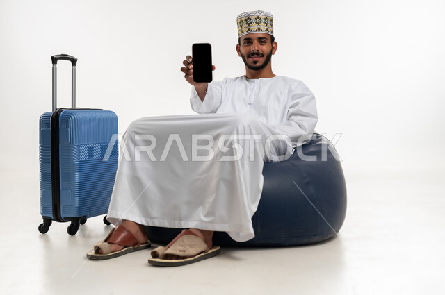 النظر الى الكاميرا بتعابير السعادة والسرور، استخدام التكنولوجيا و التقنيات الحديثة المتطورة، بورتريه  لشاب عربي خليجي عماني يرتدي الدشداشه والكمه يجلس على كرسي مريح بجانبه حقيبة سفر، عرض شاشة سوداء فارغة عبر الهاتف المحمول، خلفية بيضاء