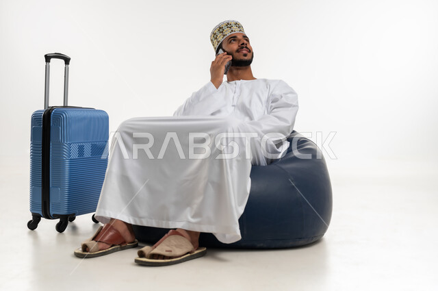 إجراء مكالمة هاتفية عبر الهاتف المحمول بتعابير الاندماج والتركيز على شيء ما، مكالمات ودردشات مع الأهل والأصدقاء، بورتريه  لشاب عربي خليجي عماني يرتدي الدشداشه والكمه يجلس على كرسي مريح وبجانبه حقيبة سفر، تبادل الأحاديث والأخبار الجديدة، خلفية بيضاء
