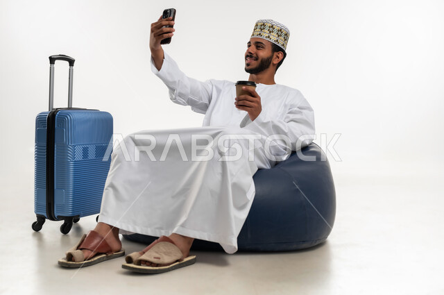 القيام بالجولات الترفيهية وقت الإجازة، مفهوم السياحة والسفر، إجراء مكالمة فيديو بايماءات لسعادة والسرور، بورتريه لشاب عربي خليجي عماني يرتدي الدشداشه والكمه يجلس على كرسي مريح يحمل بيده كأس القهوة وبجانبه حقيبة سفر، التقاط صورة سيلفي تذكارية، خلفية بيضاء