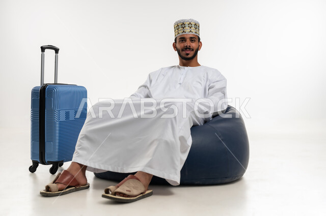 الاستعداد لرحلة السفر، بورتريه  لشاب عربي خليجي عماني يرتدي الدشداشه والكمه  يجلس على كرسي مريح وبجانبه حقيبة سفر، النظر الى الكاميرا بايماءات السعادة، قضاء رحلة ترفيهية ممتعة وقت الاجازة، خلفية بيضاء