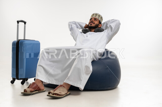 الاستمتاع بوقت الاستراحة والاسترخاء، ايماءات التركيز والنظر الى شيء ما، الاستعداد لرحلة السفر، بورتريه  لشاب عربي خليجي عماني يرتدي الدشداشه والكمه ويضع سماعة الهيدفون يجلس على كرسي مريح وبجانبه حقيبة سفر، قضاء رحلة ترفيهية ممتعة وقت الاجازة، خلفية بيضاء