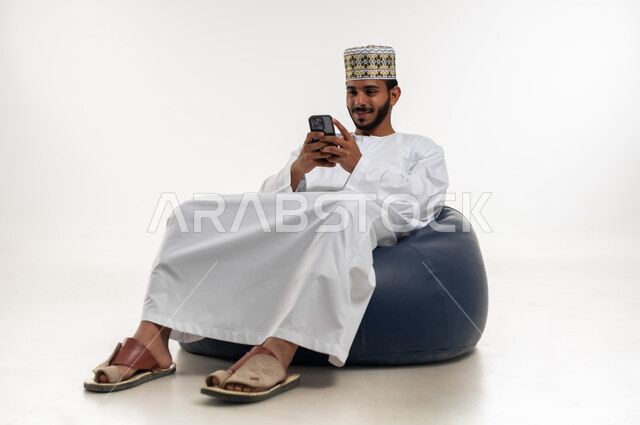 التحدث والدردشة مع الاصدقاء عبر الانترنت، تصفح تطبيقات الهاتف المحمول، استخدام التكنولوجيا و التقنيات الحديثة المتطورة، بورتريه  لشاب عربي خليجي عماني يرتدي الدشداشه والكمه يجلس على كرسي مريح يقوم بمتابعة اخبار وسائل التواصل الاجتماعي، خلفية بيضاء