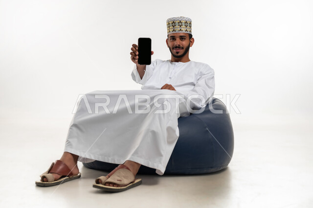 استخدام التكنولوجيا و التقنيات الحديثة المتطورة، بورتريه  لشاب عربي خليجي عماني يرتدي الدشداشه والكمه يجلس على كرسي مريح يقوم بعرض شاشة سوداء فارغة عبر الهاتف المحمول، النظر الى الكاميرا بتعابير السعادة والسرور، خلفية بيضاء