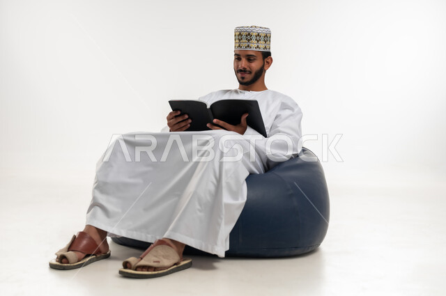 تمضية أوقات الفراغ بالتعلم والتثقف، الاهتمام بتصفح الكتب المفيدة، حب المطالعة والقراءة، بورتريه  لشاب عربي خليجي عماني يرتدي الدشداشه والكمه يجلس على كرسي مريح ينظر الى الكتاب بتمعن وتركيز، ثقافة التعليم الذاتي، خلفية بيضاء