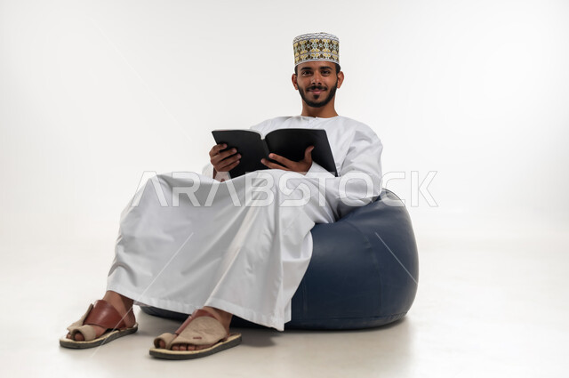 حب المطالعة والقراءة، بورتريه  لشاب عربي خليجي عماني  يرتدي الدشداشه والكمه يجلس على كرسي مريح يحمل بيده كتاب، النظر الى الكاميرا بتعابير السعادة، تمضية أوقات الفراغ بالتعلم والتثقف، الاهتمام بتصفح الكتب المفيدة، خلفية بيضاء