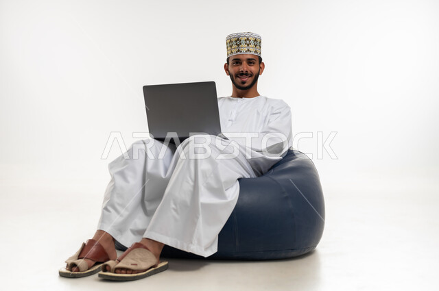 إنجاز مهام العمل عن بعد، بورتريه  لشاب عربي خليجي عماني  يرتدي الدشداشه والكمه يجلس على كرسي مريح يقوم  باستخدام الحاسوب المحمول، النظر الى الكاميرا بتعابير السرور، مفهوم التقنيات والتكنولوجيا الحديثة المتطورة، خلفية بيضاء