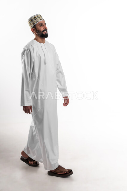 مفهوم التقدم الى الامام، بورتريه لشاب عربي خليجي عماني مبتسم يرتدي الدشداشة و الكمه ينظر الى الامام بإيماءات تدل على الحركة والمشي، الشعور بالسرور والفرح، صورة الجسم بالطول الكامل, خلفية بيضاء