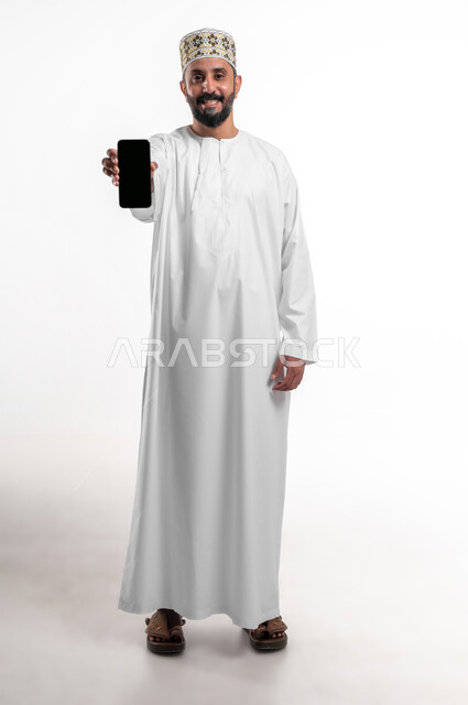تعابير السعادة والفرح، استخدام الأجهزة والتقنيات الحديثة، بورتريه مقرب لشاب عربي خليجي عماني يرتدي الدشداشة و الكمه يمسك بيده الهاتف المحمول يقوم بعرض شاشة سوداء فارغة بإيماءات السرور، صورة الجسم بالطول الكامل, خلفية بيضاء