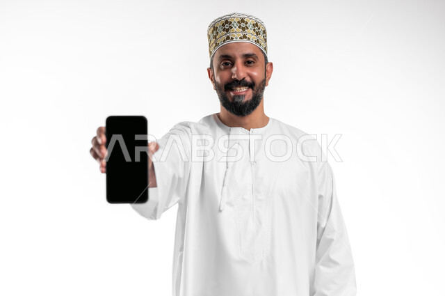 تعابير السعادة والفرح، استخدام الأجهزة والتقنيات الحديثة، بورتريه مقرب لشاب عربي خليجي عماني يرتدي الدشداشة و الكمه يمسك بيده الهاتف المحمول يقوم بعرض شاشة سوداء فارغة بإيماءات السرور، خلفية بيضاء