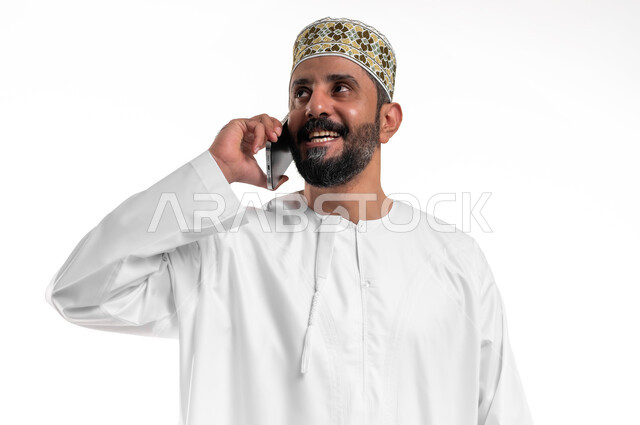 الدردشة والتواصل مع الاصدقاء بإيماءات السعادة والفرح، استخدام الأجهزة والتقنيات الحديثة، بورتريه مقرب لشاب عربي خليجي عماني يرتدي الدشداشة و الكمه يمسك بيده الهاتف المحمول يقوم بإجراء المكالمات الهاتفية، خلفية بيضاء