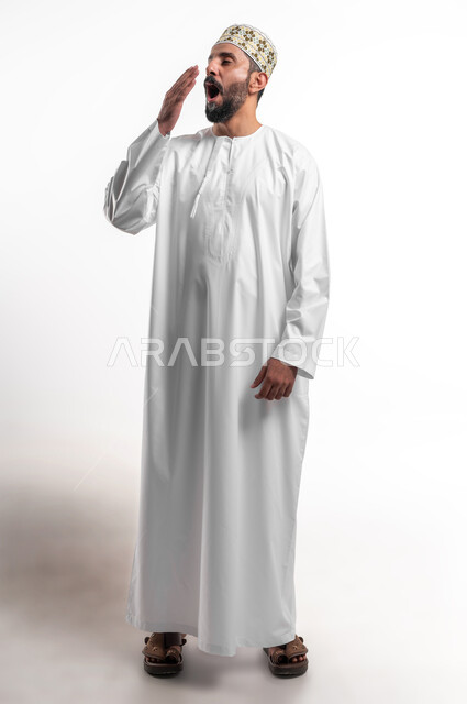 الشعور بالتعب والحاجة الى النوم والراحة, الإحساس بالكسل والخمول و الإرهاق، بورتريه مقرب لشاب عربي خليجي عماني مبتسم يرتدي الدشداشة و الكمه يضع يده على فمه بإيماءات التثاؤب والنعاس، صورة الجسم بالطول الكامل, خلفية بيضاء
