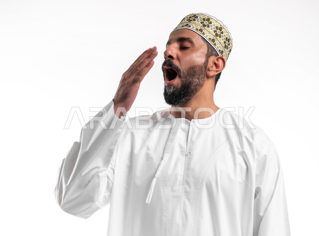 الشعور بالتعب والحاجة الى النوم والراحة, الإحساس بالكسل والخمول و الإرهاق، بورتريه مقرب لشاب عربي خليجي عماني مبتسم يرتدي الدشداشة و الكمه يضع يده على فمه بإيماءات التثاؤب والنعاس، خلفية بيضاء