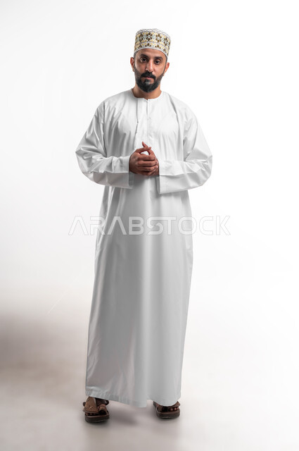 Solving problems and making decisions, feeling upset and bored, a close-up portrait of a young Arab Gulf Omani man wearing a dishdasha and a cuff, looking at the camera with gestures of seriousness and annoyance, the concept of thinking and bewilderment, full-length body portrait, white background