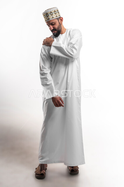 الأوجاع والآلام العصبية والعضلية، طلب المساعده والحاجه للعلاج، التهاب العضلات والمفاصل، بورتريه لشاب عربي خليجي عماني يرتدي الدشداشة و الكمه يضع يده على كتفه بايماءات الألم والتعب والأرهاق، حالة اسعافية طارئة، صورة الجسم الطول الكامل, خلفية بيضاء