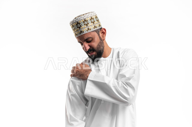 الأوجاع والآلام العصبية والعضلية، طلب المساعده والحاجه للعلاج، التهاب العضلات والمفاصل، بورتريه لشاب عربي خليجي عماني يرتدي الدشداشة و الكمه يضع يده على كتفه بايماءات الألم والتعب والأرهاق، حالة اسعافية طارئة، صورة الجسم الطول الكامل, خلفية بيضاء