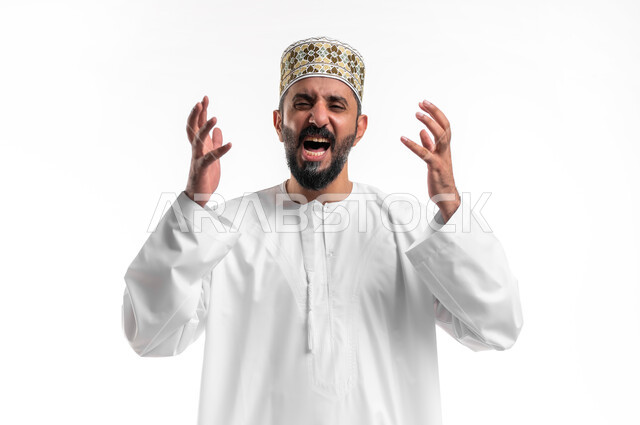 مفهوم الغضب و الإثاره والرغبة بالانتقام, بورتريه مقرب لشاب عربي خليجي عماني مبتسم يرتدي الدشداشة و الكمه يرفع يديه للاعلى بإيماءات الصراخ، الحزن والقهر من الخسارة، خلفية بيضاء
