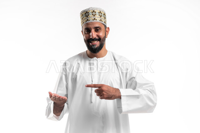 تعابير الفرح والسرور، بورتريه لشاب عربي خليجي عماني مبتسم يرتدي الدشداشة و الكمه يحمل بيده شيء ما ويشير بإصبع السبابة ليده الأخرى، الامساك بعنصر ما بإيماءات السعادة، خلفية بيضاء