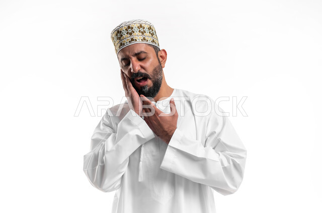 تسوس الأضراس وإلتهاب اللثه، طلب العلاج و المساعده، بورتريه مقرب لشاب عربي خليجي عماني يرتدي الدشداشة و الكمه يضع يديه على خده ويغمض عينيه بإيماءات بإيماءات تدل على الشعور بألم الأسنان، الحاجه لزيارة الطبيب، خلفية بيضاء